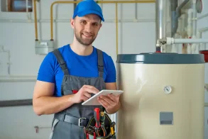 A plumber in a uniform finalizes a bill for a customer in a clean, finished basement with a new water heater installed.